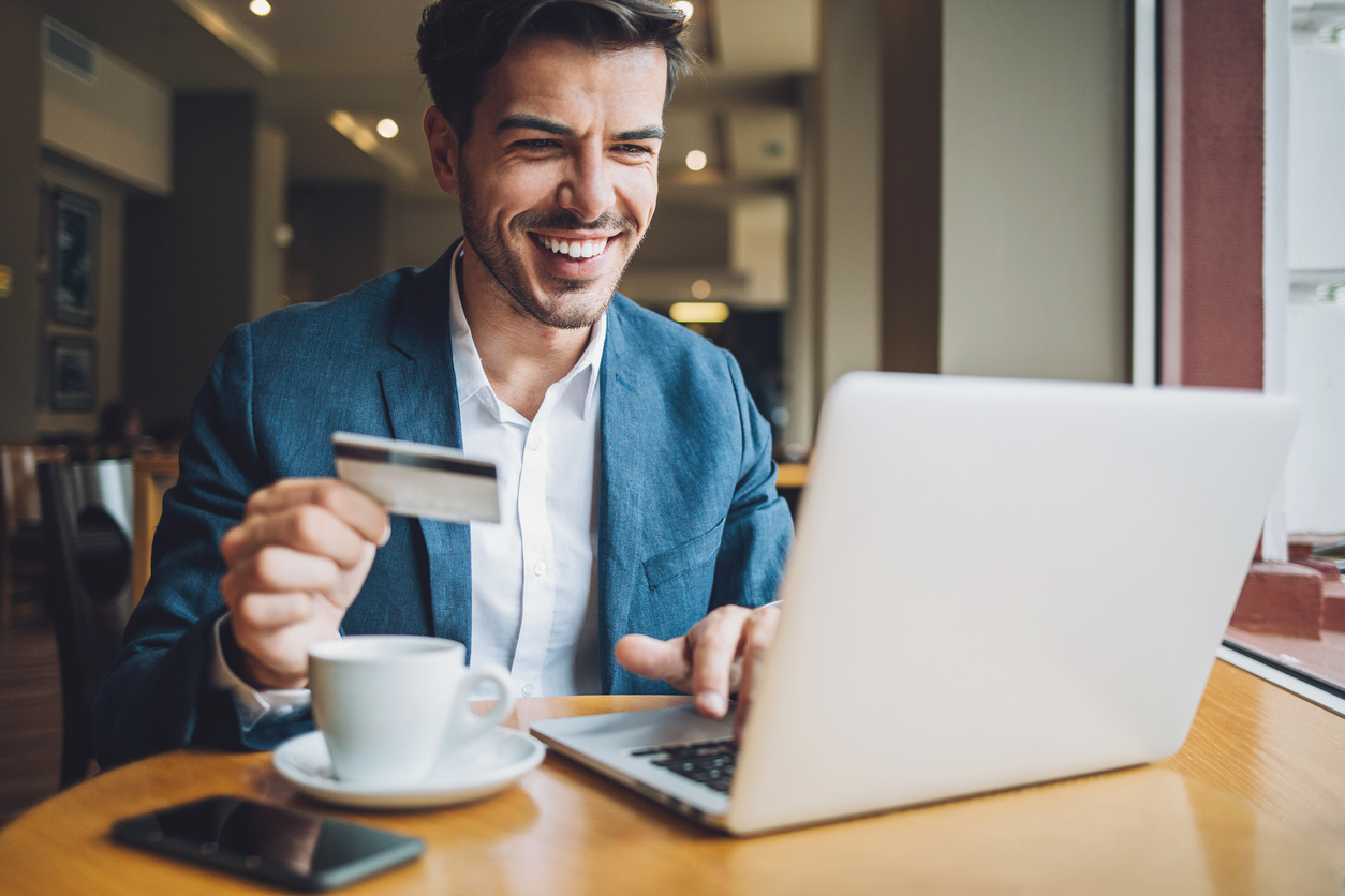 Smiling man sitting in cafe, holding a credit card and typing on a laptop.